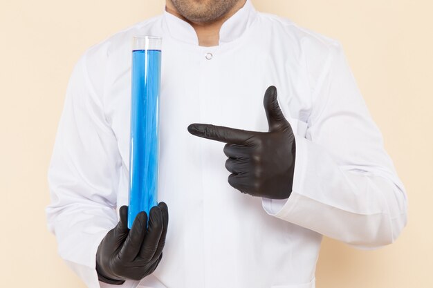 クリーム色の机の実験室の化学科学実験で青い溶液と小さなフラスコを保持している白い特別なスーツの正面図若い男性の化学者