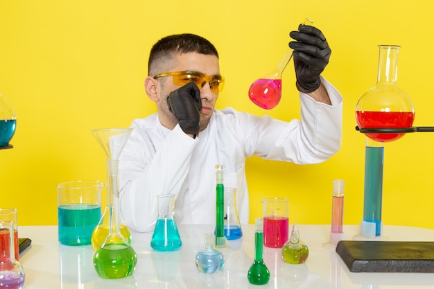 플라스크를 들고 노란색 책상 과학 작업 실험실에 생각 컬러 솔루션으로 테이블 앞에 흰색 정장에 전면보기 젊은 남성 화학자