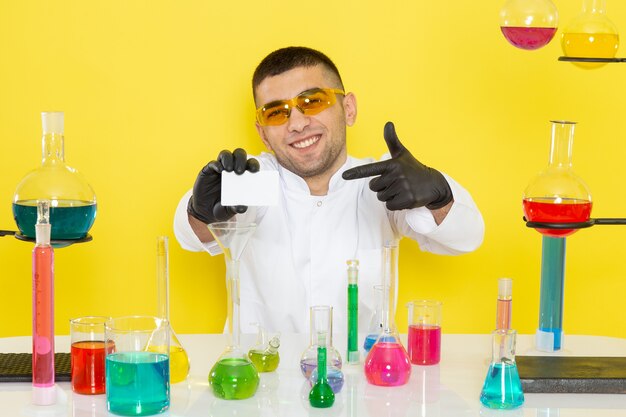Вид спереди молодой химик-мужчина в белом костюме перед столом с цветными растворами, держащий белую карточку, улыбающийся на желтой стене, научная работа, лаборатория, химия