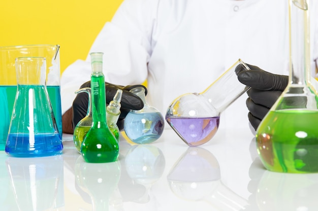 Front view young male chemist in white suit in front of table with colored solutions working with them on the yellow wall science work lab chemistry