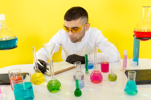 Вид спереди молодой химик в белом костюме перед столом с цветными растворами, записывающий заметки на желтой стене, научная работа, химическая лаборатория
