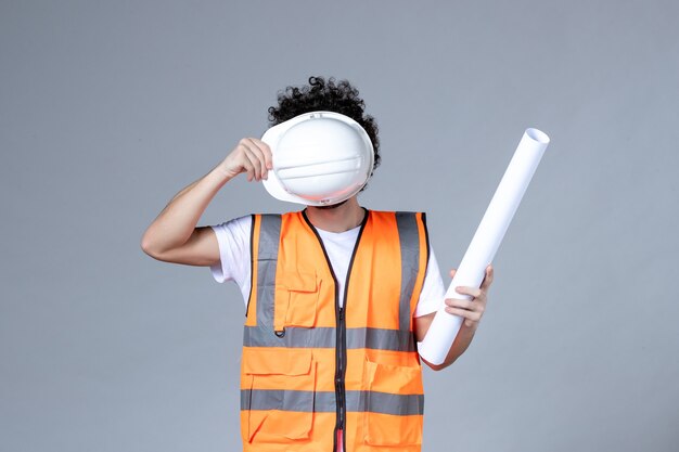 Front view of young male constructor in warning vest holding safety helmet in front of his face and showing blank on gray wall