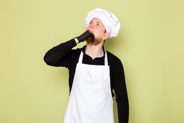 A front view young male cook in black shirt with white cape white head cap in black gloves posing thinking