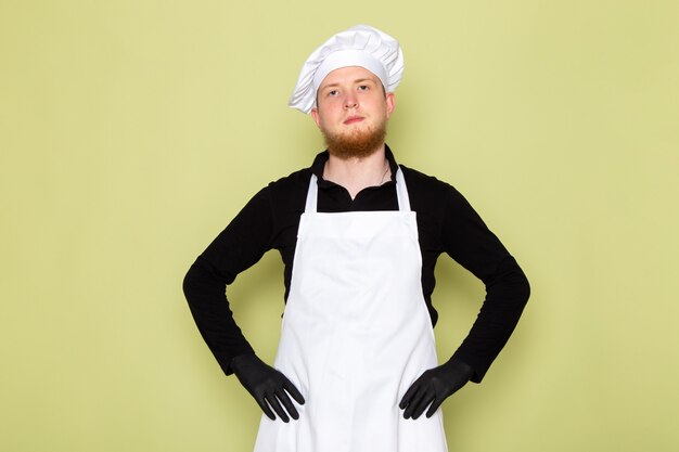 A front view young male cook in black shirt with white cape white head cap in black gloves posing