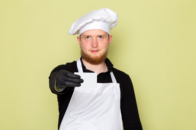 A front view young male cook in black shirt with white cape white head cap in black gloves showing grey card smiling