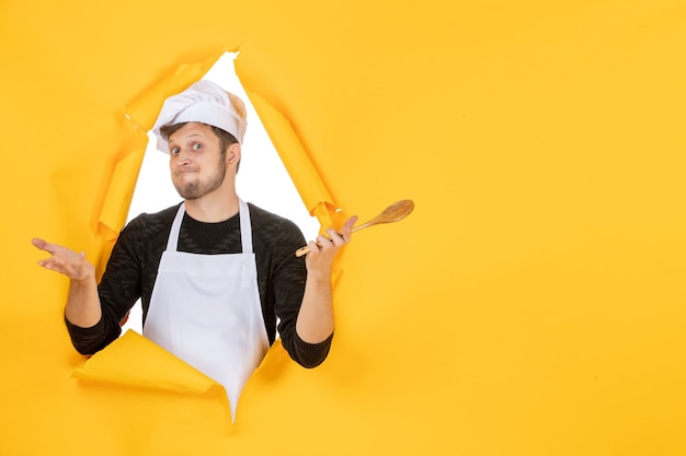 Front view young male cook in white cape holding wooden spoon on yellow background color kitchen photo cuisine job white man