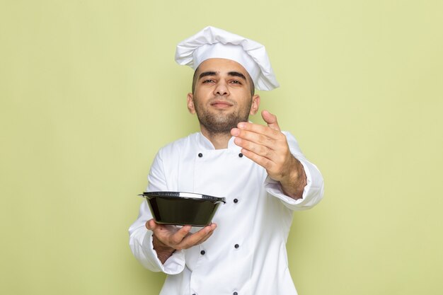 Front view young male cook in white cook suit holding black food bowl on green