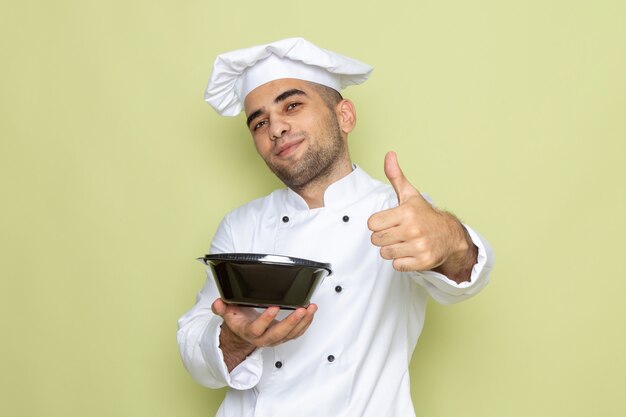 Front view young male cook in white cook suit holding black food bowl and showing like sign on green