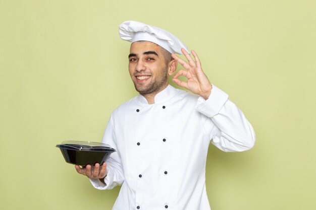 Front view young male cook in white cook suit holding black food bowl with smile on green