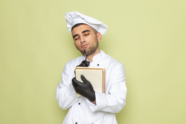 Front view young male cook in white cook suit holding notepad with thinking expression on green