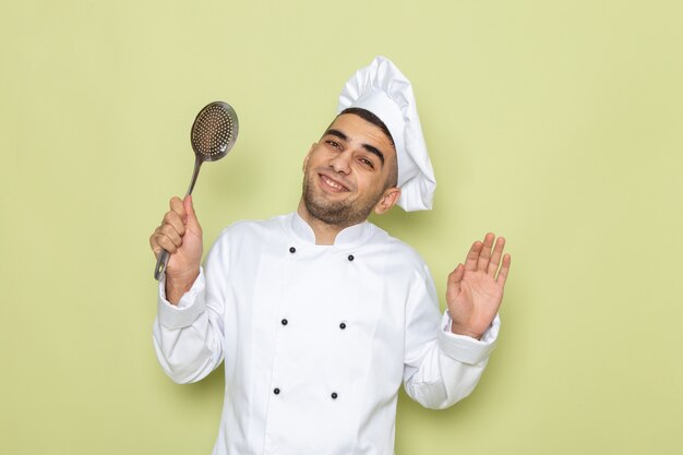 Giovane cuoco maschio di vista frontale in vestito bianco del cuoco che tiene cucchiaio d'argento con il sorriso sul verde