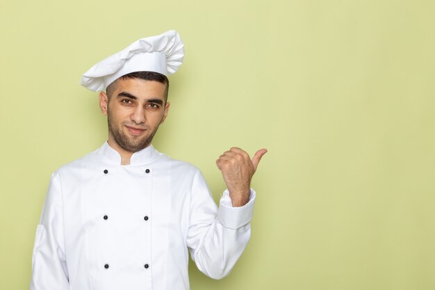 Front view young male cook in white cook suit posing with raised finger on green