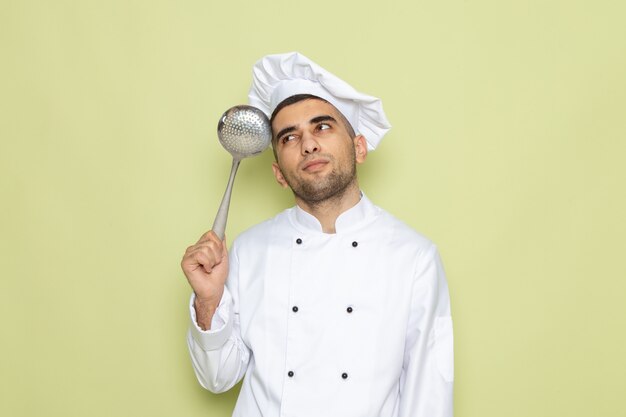 Front view young male cook in white cook suit posing with silver spoon on green