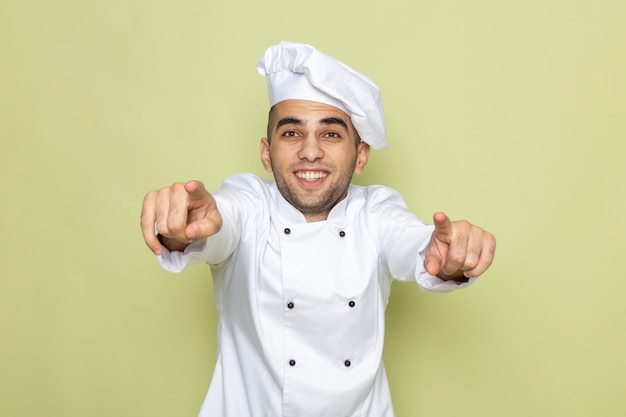 Front view young male cook in white cook suit smiling and pointing out on green