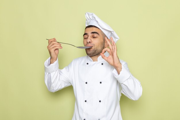 Front view young male cook in white cook suit tasting silver spoon on green