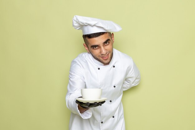 Front view young male cook in white cook suit wearing dark gloves and servicing coffee on green