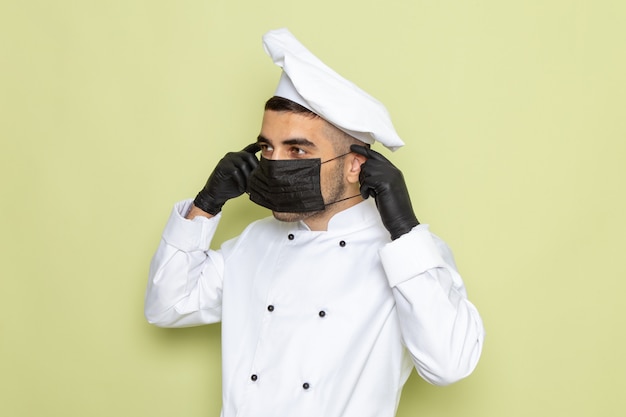 Foto gratuita giovane cuoco maschio di vista frontale in vestito bianco da cuoco che indossa guanti scuri e indossa una maschera sul maschio uniforme di cucina di lavoro verde