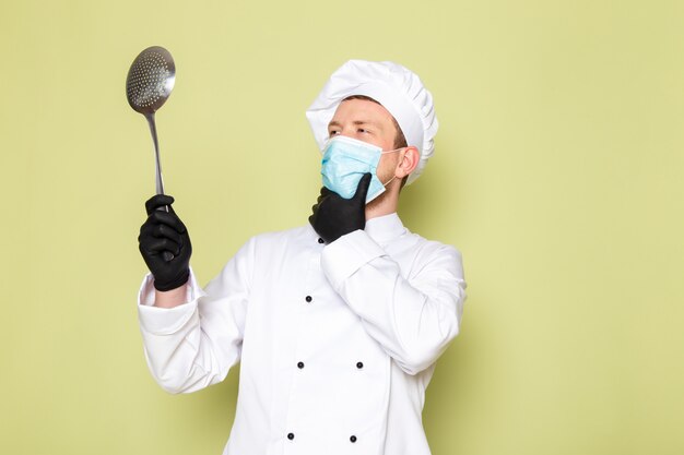 A front view young male cook in white cook suit white head cap in black gloves blue protective mask holding big metallic spoon thinking