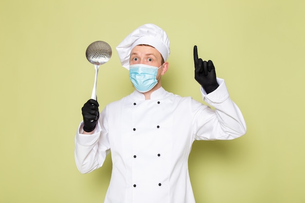 Un cuoco maschio giovane di vista frontale in cappuccio bianco bianco del vestito del cuoco in maschera protettiva blu dei guanti neri che tiene grande cucchiaio metallico