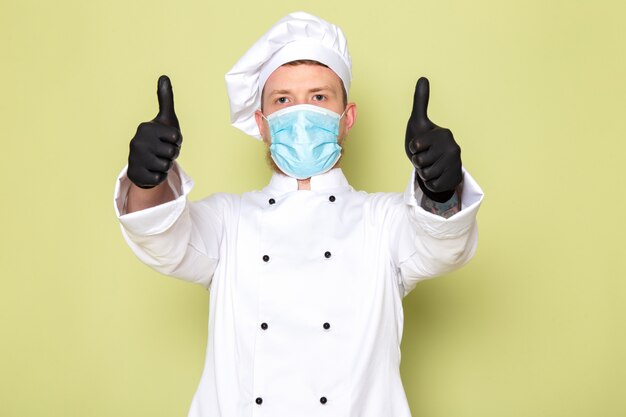 A front view young male cook in white cook suit white head cap in black gloves blue protective mask showing awesome sign