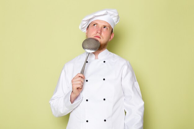 Вид спереди молодой мужчина повар в белом костюме повара белая шапка держит большую серебряную ложку, думая