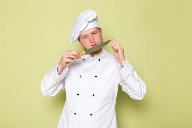 A front view young male cook in white cook suit white head cap holding checking knife