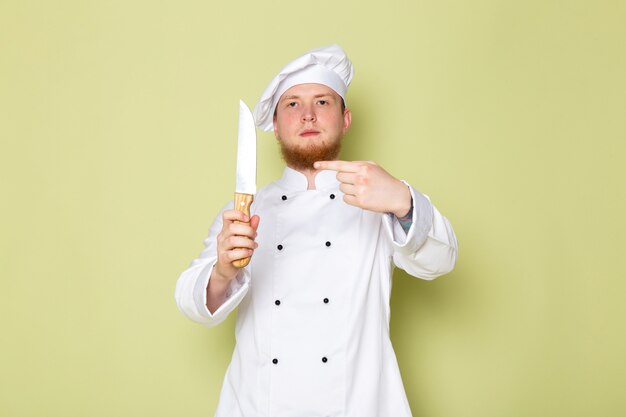 A front view young male cook in white cook suit white head cap holding knife