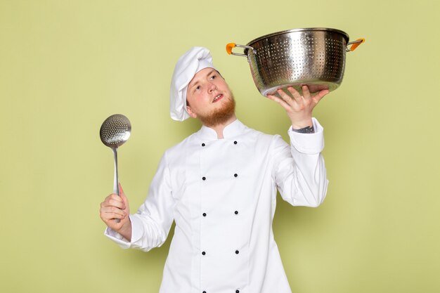 흰색 쿡 정장 흰색 머리 모자 큰 은색 스푼으로 은색과 금속 냄비를 들고 전면보기 젊은 남성 요리사