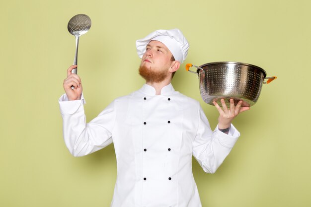 A front view young male cook in white cook suit white head cap holding silver and metallic saucepan with big silver spoon