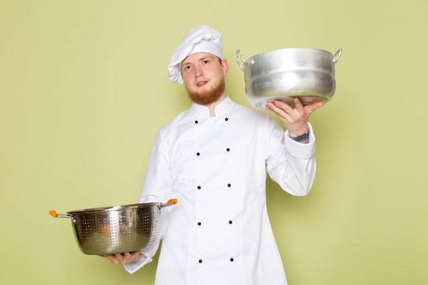 A front view young male cook in white cook suit white head cap holding silver and metallic saucepans