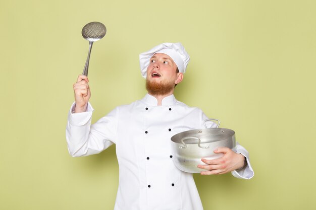 A front view young male cook in white cook suit white head cap holding silver saucepan and big silver spoon