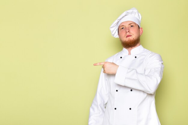 A front view young male cook in white cook suit white head cap pointing