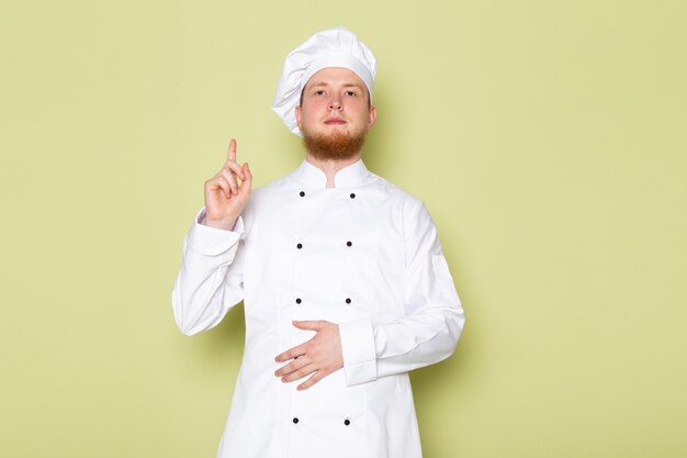 A front view young male cook in white cook suit white head cap raising his finger