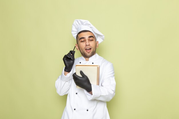 Front view young male cook in white cook suit writing down notes with gloves on green