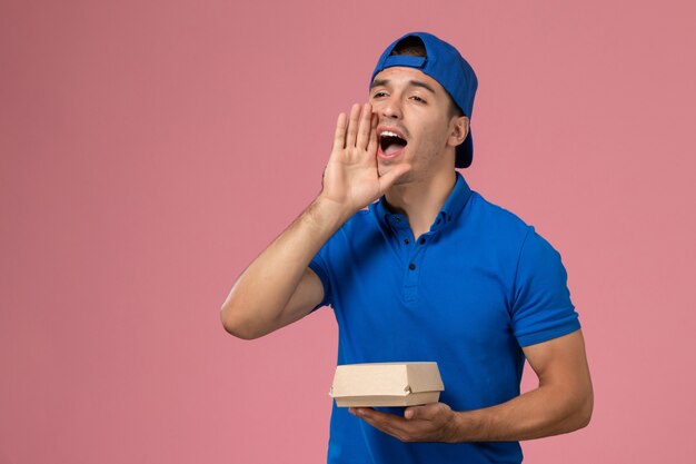 Front view young male courier in blue uniform cape holding delivery food package calling out on pink wall