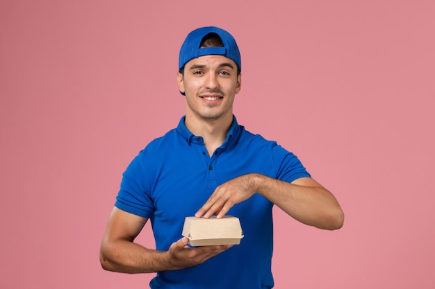 ピンクの壁に配達食品パッケージを保持している青い制服ケープの正面図若い男性宅配便