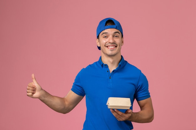 Front view young male courier in blue uniform cape holding delivery food package on pink wall