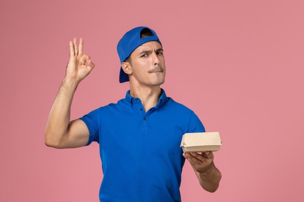 Front view young male courier in blue uniform cape holding delivery food package on pink wall