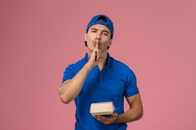 Front view young male courier in blue uniform cape holding delivery food package showing silence sign on pink wall