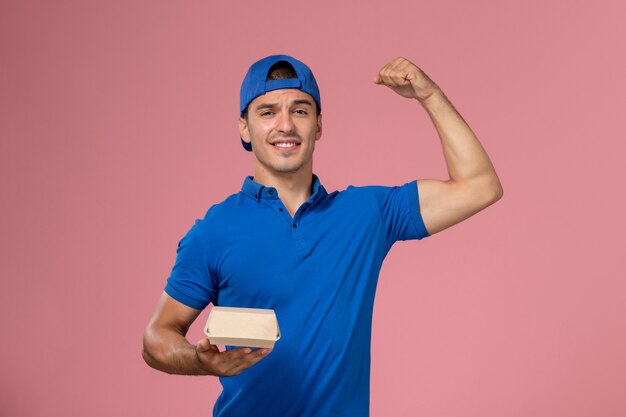 Front view young male courier in blue uniform cape holding little delivery food package flexing on the pink wall