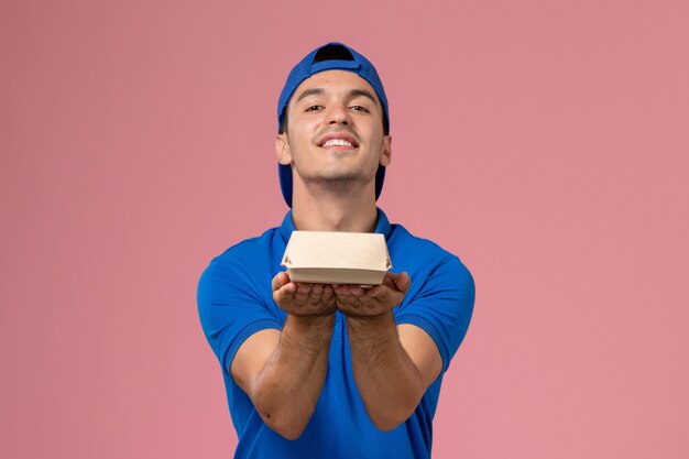 Front view young male courier in blue uniform cape holding little delivery food package on the light-pink wall
