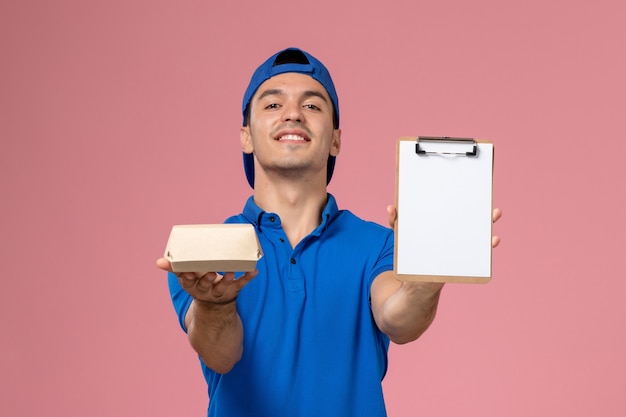 Front view young male courier in blue uniform cape holding little delivery food package and notepad on the light-pink wall