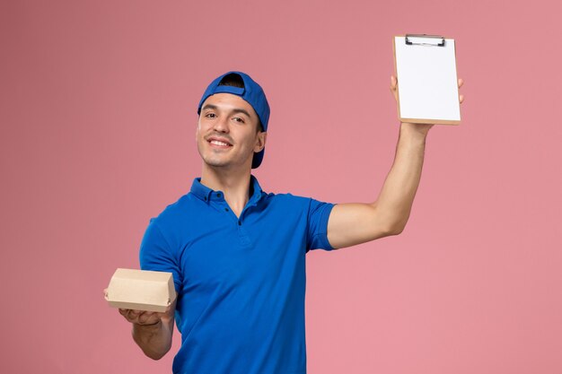 Front view young male courier in blue uniform cape holding little delivery food package and notepad on the light-pink wall