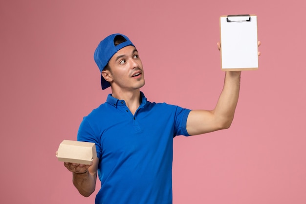 Front view young male courier in blue uniform cape holding little delivery food package and notepad on the light-pink wall