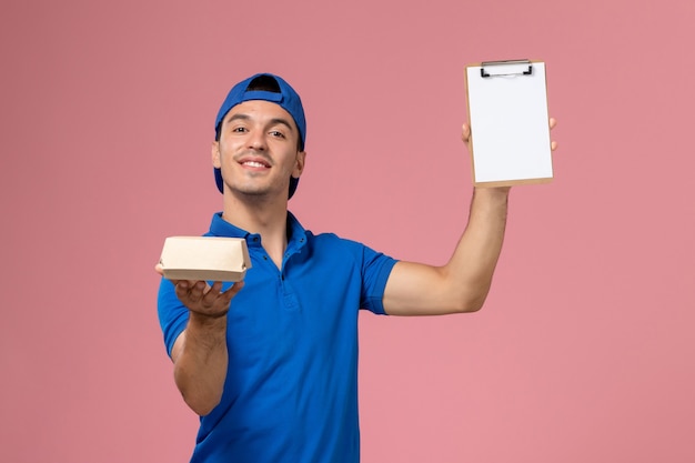 Front view young male courier in blue uniform cape holding little delivery food package and notepad on the light-pink wall