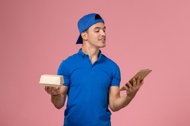 Giovane corriere maschio di vista frontale in capo uniforme blu che tiene poco pacchetto dell'alimento di consegna e blocchetto per appunti sulla parete rosa-chiaro