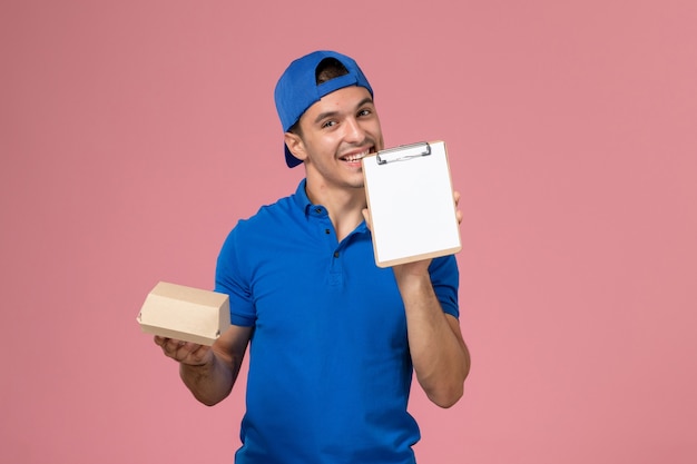 Front view young male courier in blue uniform cape holding little delivery food package and notepad on the light-pink wall