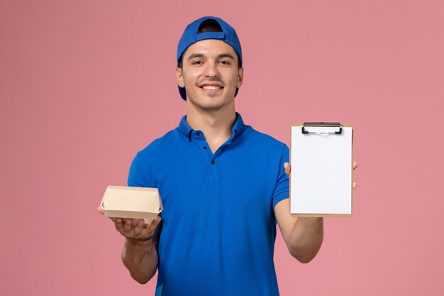 Front view young male courier in blue uniform cape holding little delivery food package and notepad smiling on the light-pink wall