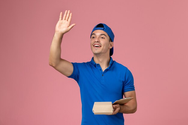 Front view young male courier in blue uniform cape holding little delivery food package and notepad waving on light-pink wall