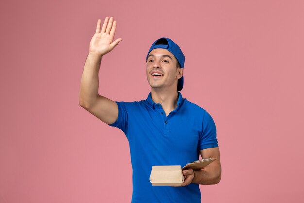 淡いピンクの壁に手を振っている小さな配達食品パッケージとメモ帳を保持している青い制服の岬の正面図若い男性の宅配便
